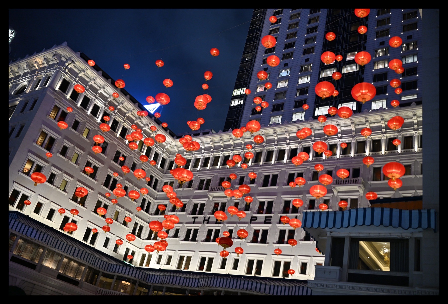 夜色庭院上方悬挂着密集红灯笼，旧建筑与高层立面围合出向上的视线
