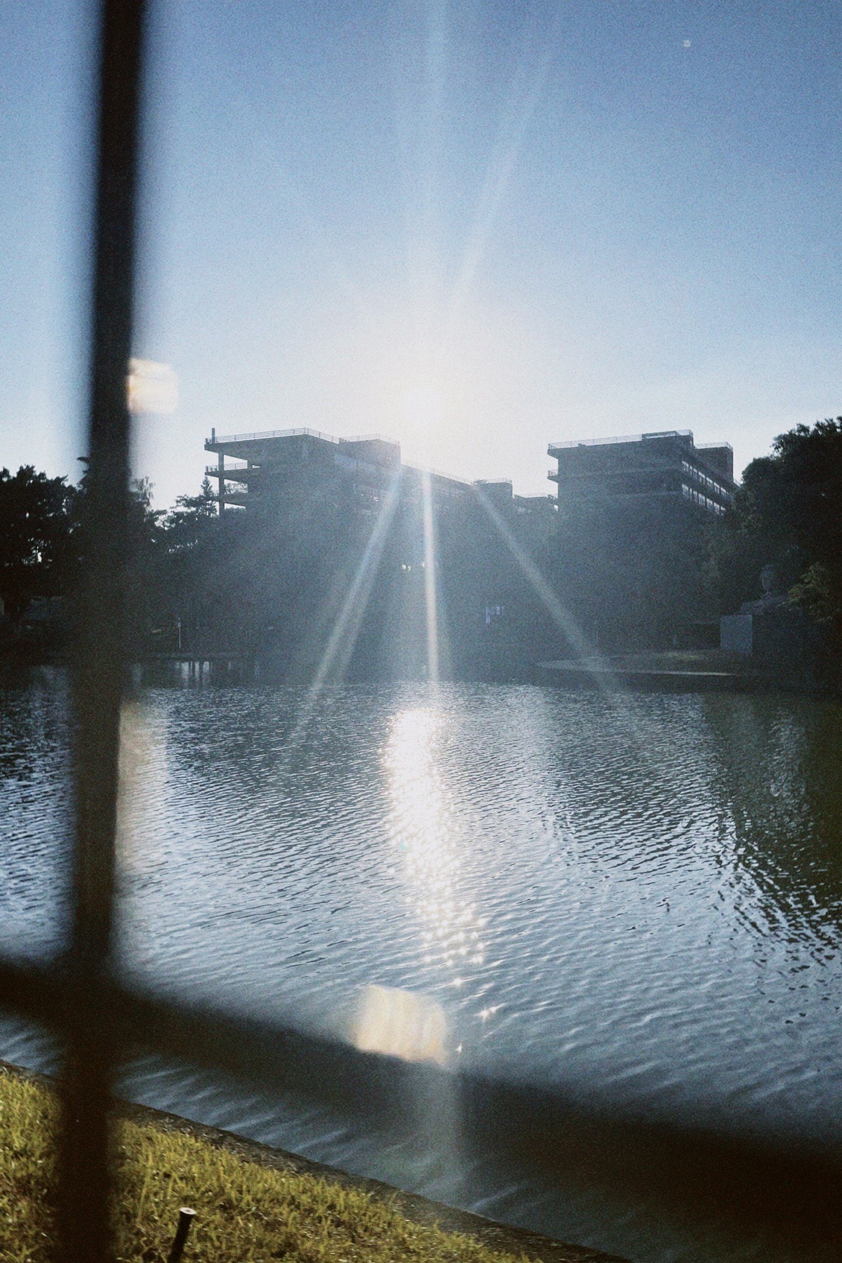 隔着暗色栏杆望向湖面，逆光在水面和建筑轮廓间形成垂直光束